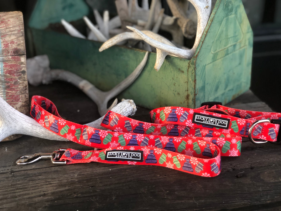 Red winter-themed Lucky Love Dog collar and leash set with mitten and hat pattern displayed on rustic wood with antler decor.