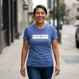 A woman smiling while walking on a city sidewalk, wearing a heathered blue crewneck t-shirt with a white rectangular chest graphic that reads 'LUCKY I LOVE DOG AUSTIN TX' and smaller text below.