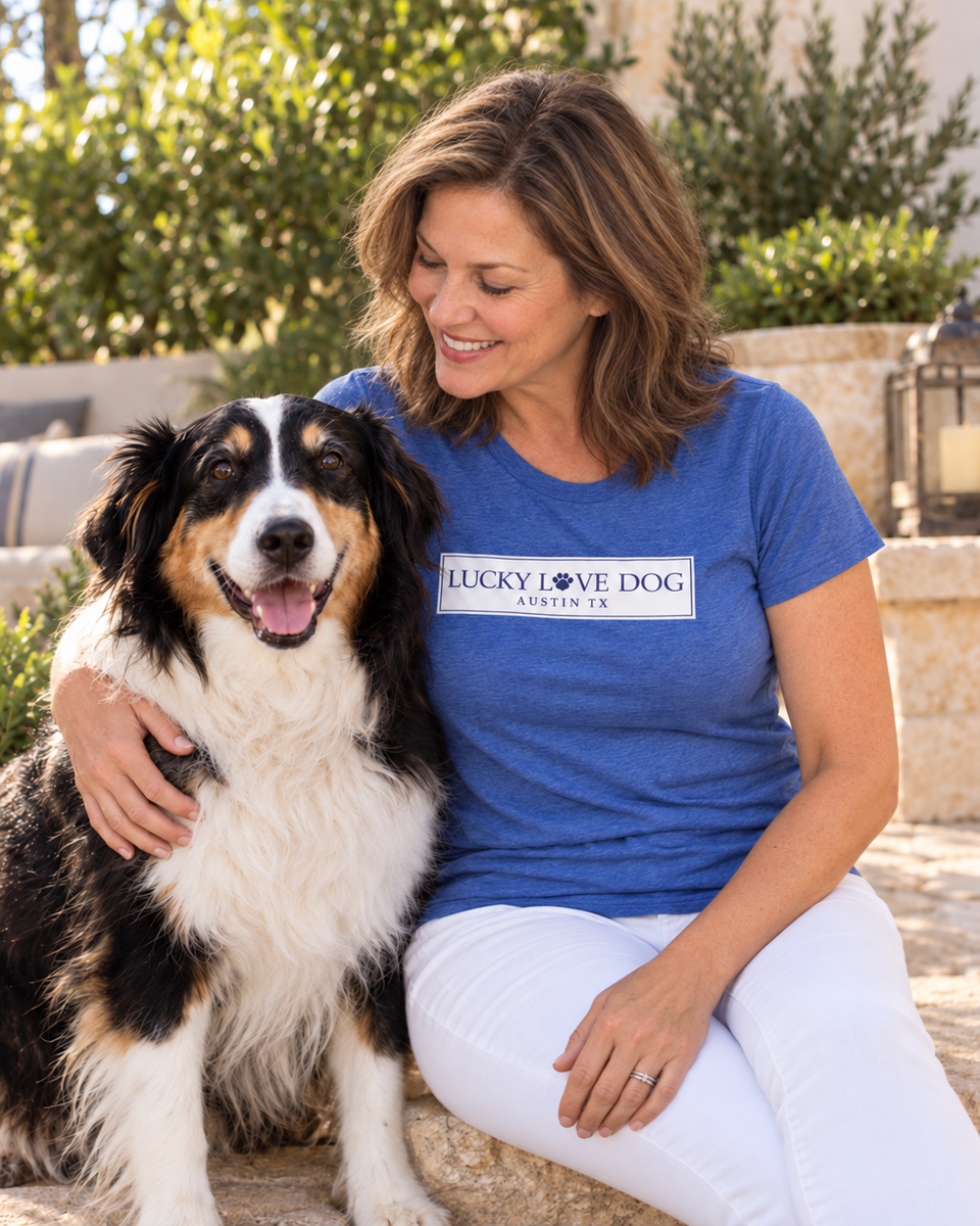 A woman sitting outdoors next to a tricolor Australian Shepherd dog, wearing a blue t-shirt with a white graphic that reads 'LUCKY LOVE DOG - DENMARK'.