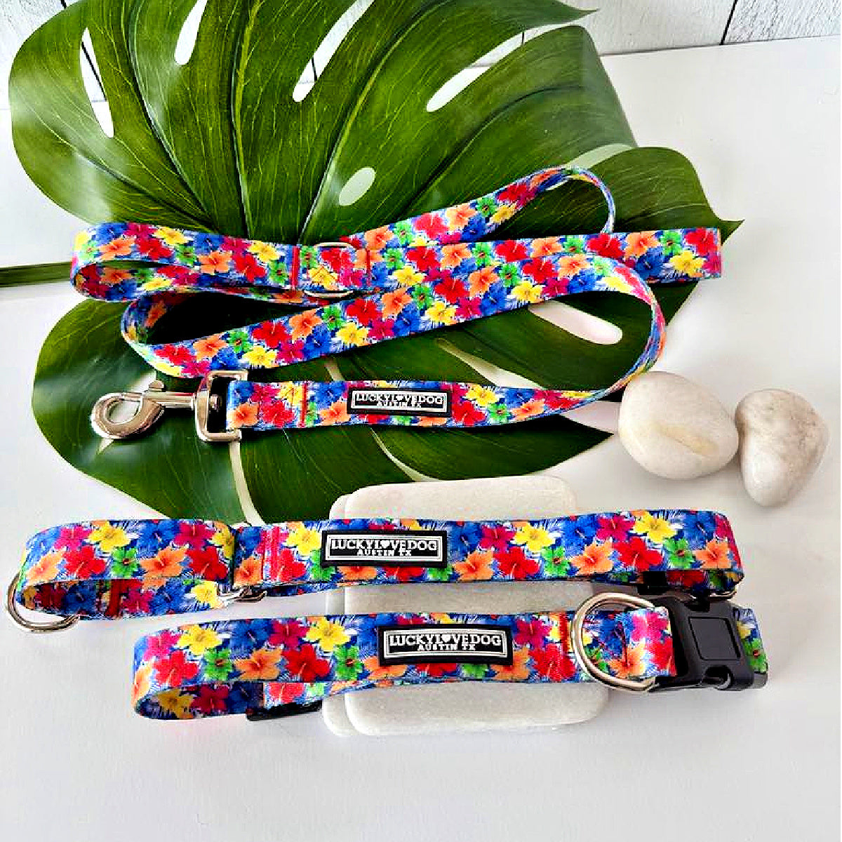 Tropical hibiscus patterned dog leash, martingale, and standard collar set displayed on rocks and a large green leaf.