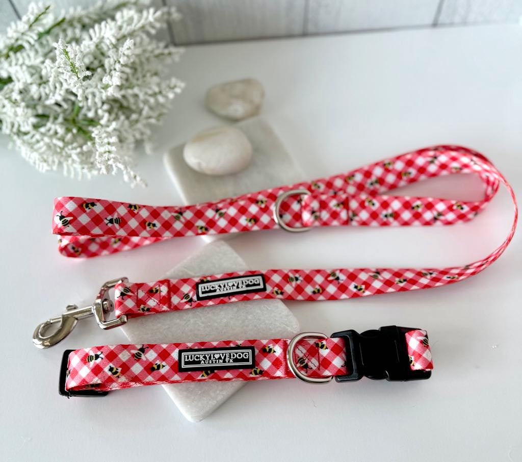 Red gingham dog collar, martingale, and leash set with a bumblebee pattern, displayed on marble with flowers.