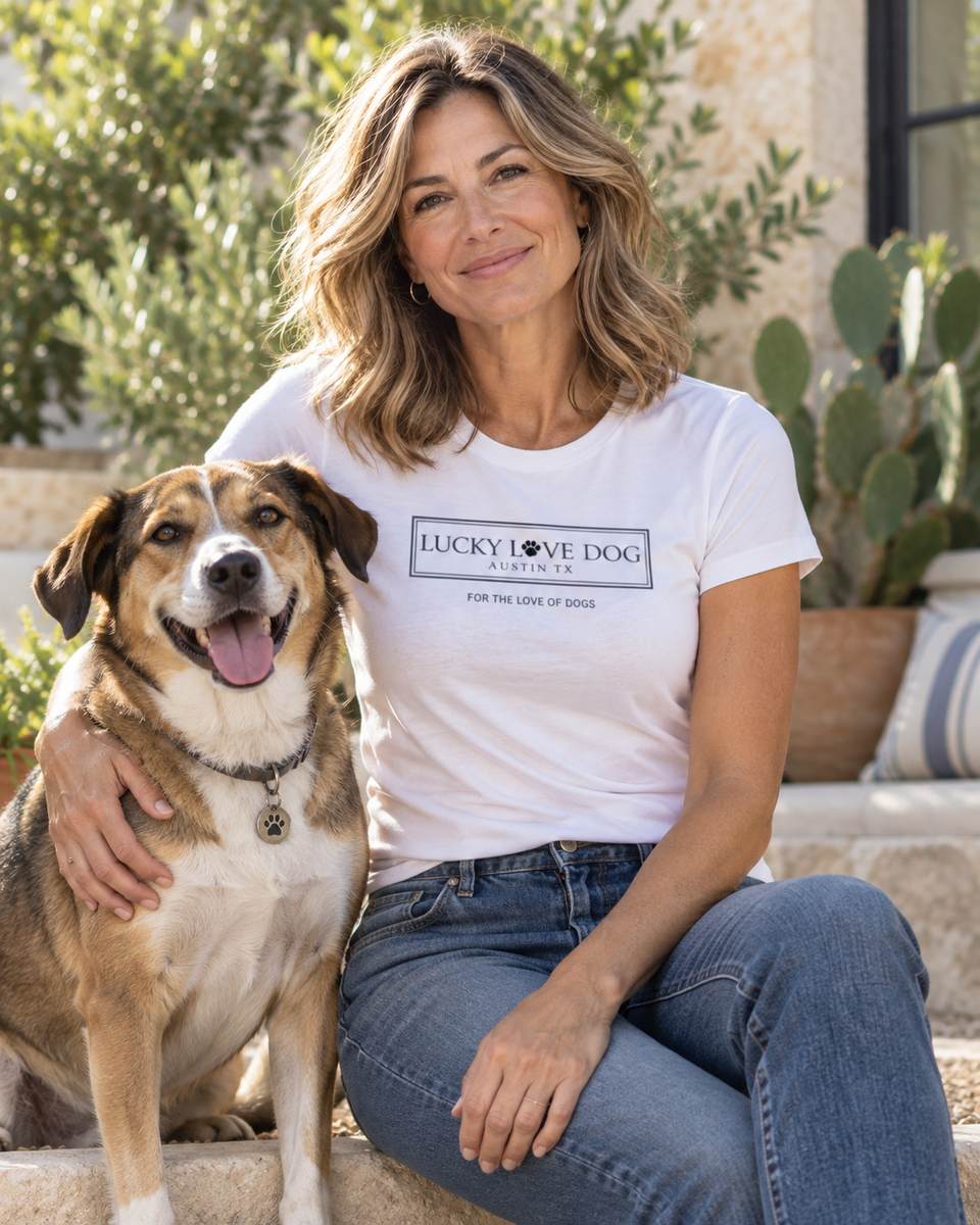 A woman sitting outdoors next to a brown and white dog, wearing a white graphic t-shirt that reads 'LUCKY LOVE DOG PET SUPPLIES' and 'FOR THE LOVE OF DOGS'.