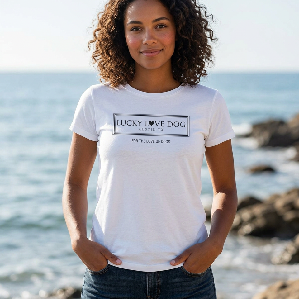 A woman with curly brown hair stands on a rocky beach wearing a white short-sleeve graphic T-shirt that reads 'LUCKY DOG AUSTIN TX' and 'FOR THE LOVE OF DOGS' in black text. The background shows the ocean and rocks in soft focus.