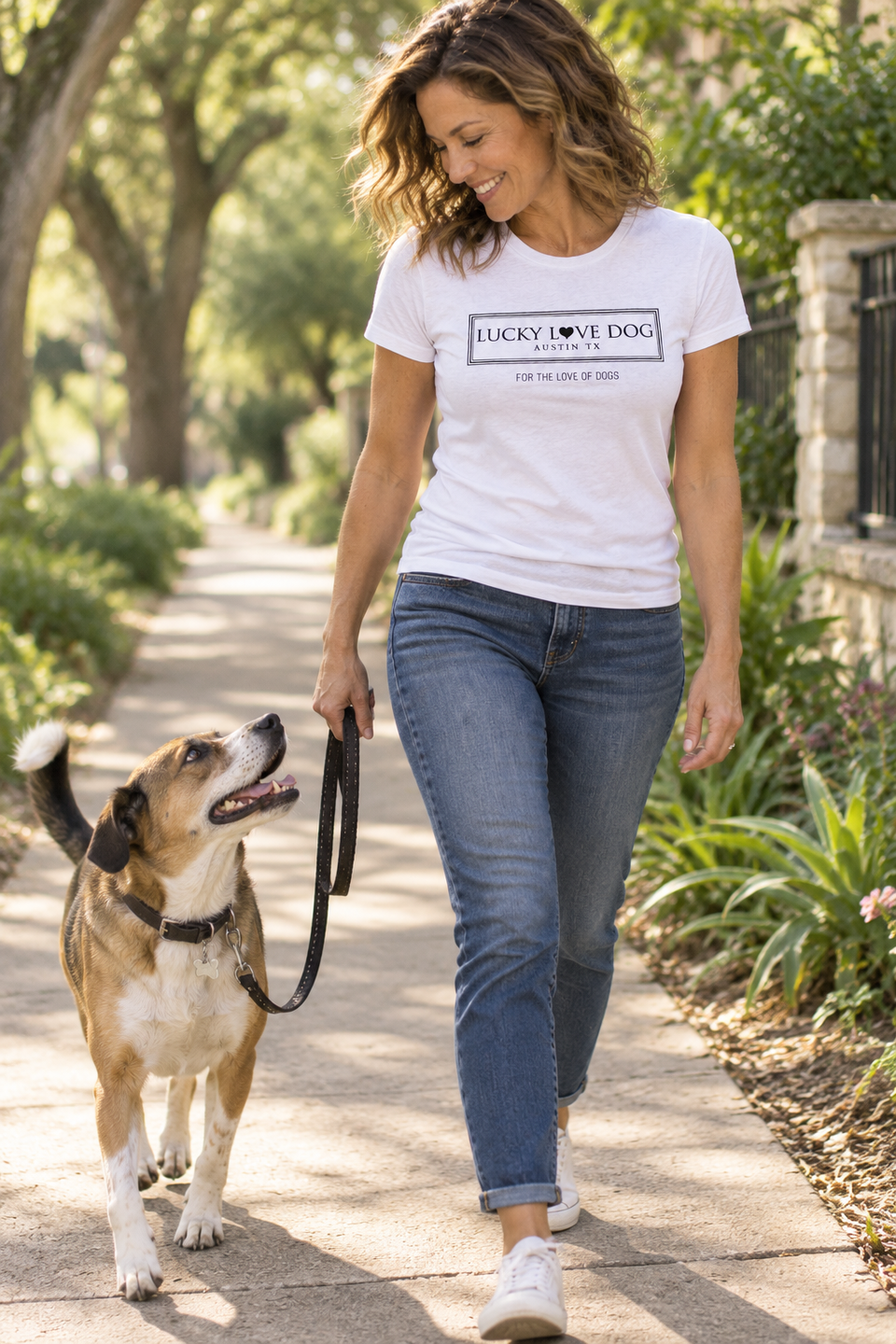 A woman walking a dog on a leash along a sunlit suburban path. She is wearing a white graphic T-shirt with the text 'LUCKY DOG PET POLICE OFFICE' and blue jeans.