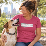 A woman sitting outdoors with a dog, wearing a pink t-shirt with a white graphic that reads 'LUCKY LOVE DOG AUSTIN TX FOR THE LOVE OF DOGS'.