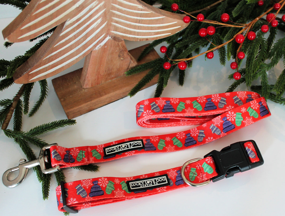 Red winter-themed Lucky Love Dog collar and leash set with mittens and hats displayed alongside festive holiday greenery.