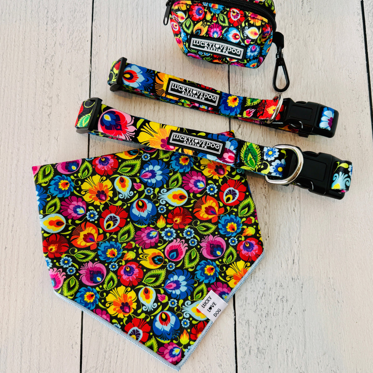 Floral dog bandana featuring a vibrant multicolored folk art pattern, displayed on a wooden surface alongside matching pet collars and a small accessory pouch.