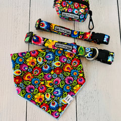 Floral dog bandana featuring a vibrant multicolored folk art pattern, displayed on a wooden surface alongside matching pet collars and a small accessory pouch.