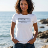 A woman with curly brown hair stands on a rocky beach wearing a white short-sleeve graphic T-shirt that reads 'LUCKY DOG AUSTIN TX' and 'FOR THE LOVE OF DOGS' in black text. The background shows the ocean and rocks in soft focus.